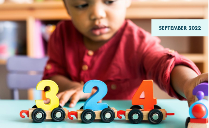 A Black toddler in a red shirt plays with a toy train in a day care center.