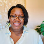 Ebony Benzing smiles for her headshot. She wears glasses and a light blue button-down shirt.