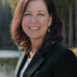 Professional shot of Shannon Moran. She wears a dark suit and stands outside with a backdrop of trees and a lake.