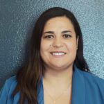 Headshot of Patricia Salinas. She has dark hair and wears a blue shirt against a lighter blue background.