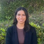 Headshot of Liza Chavoc. She is outside with plants in the background. She wears a dark blazer and is smiling.