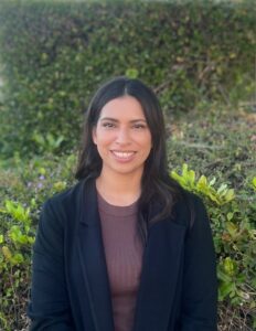 Headshot of Liza Chavoc. She is outside with plants in the background. She wears a dark blazer and is smiling.