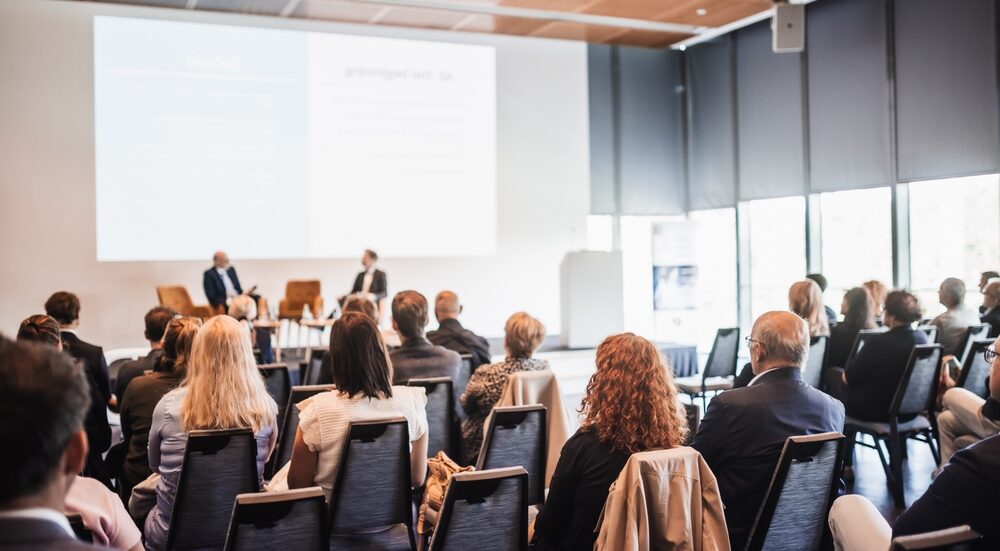An audience sits in a well-lit room, watching two presenters on a low stage.